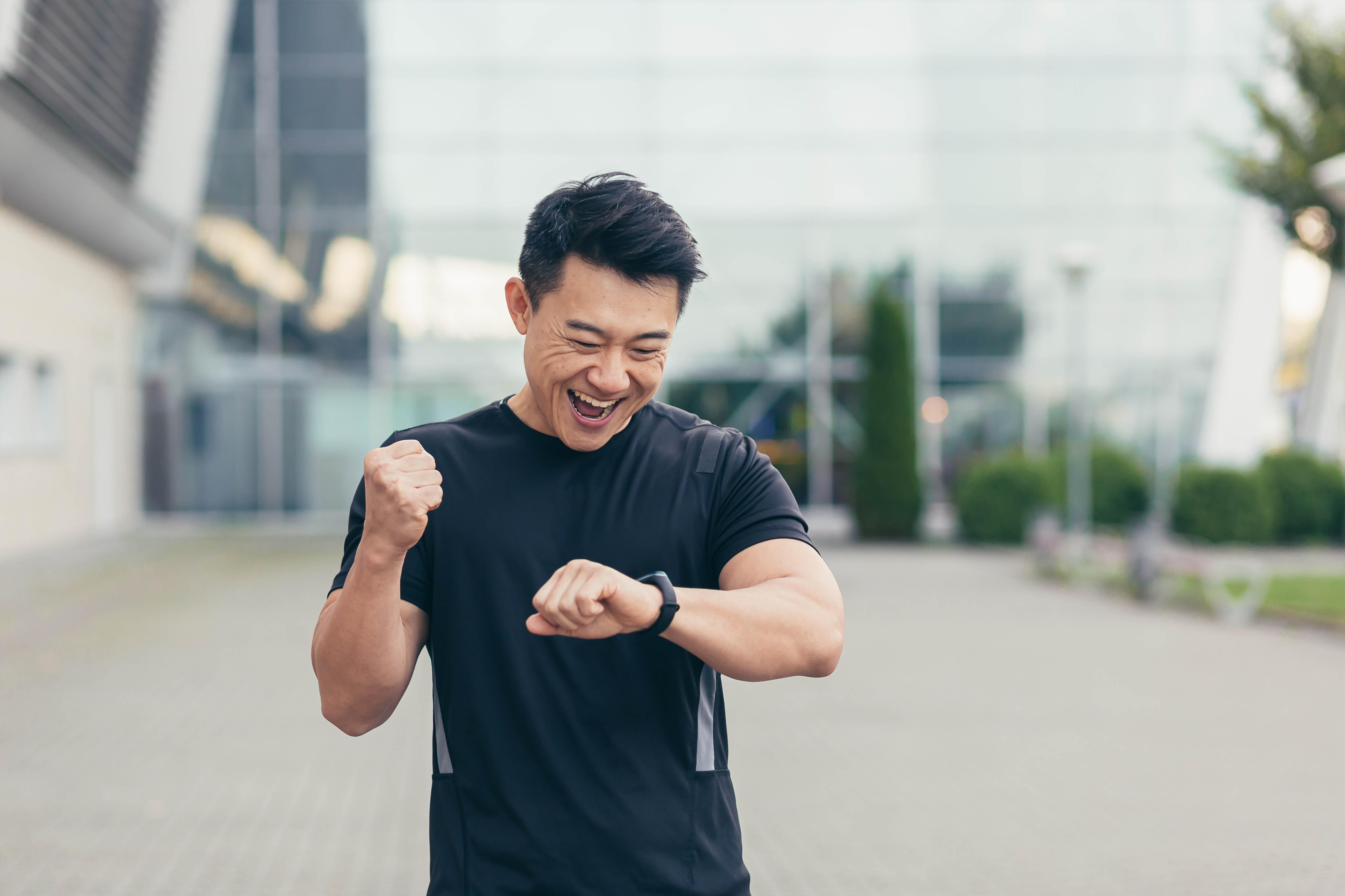Excited young Asian man looking at his fitness watch and throwing his fist in the air in excitement Excited young Asian man looking at his fitness watch and throwing his fist in the air in excitement
